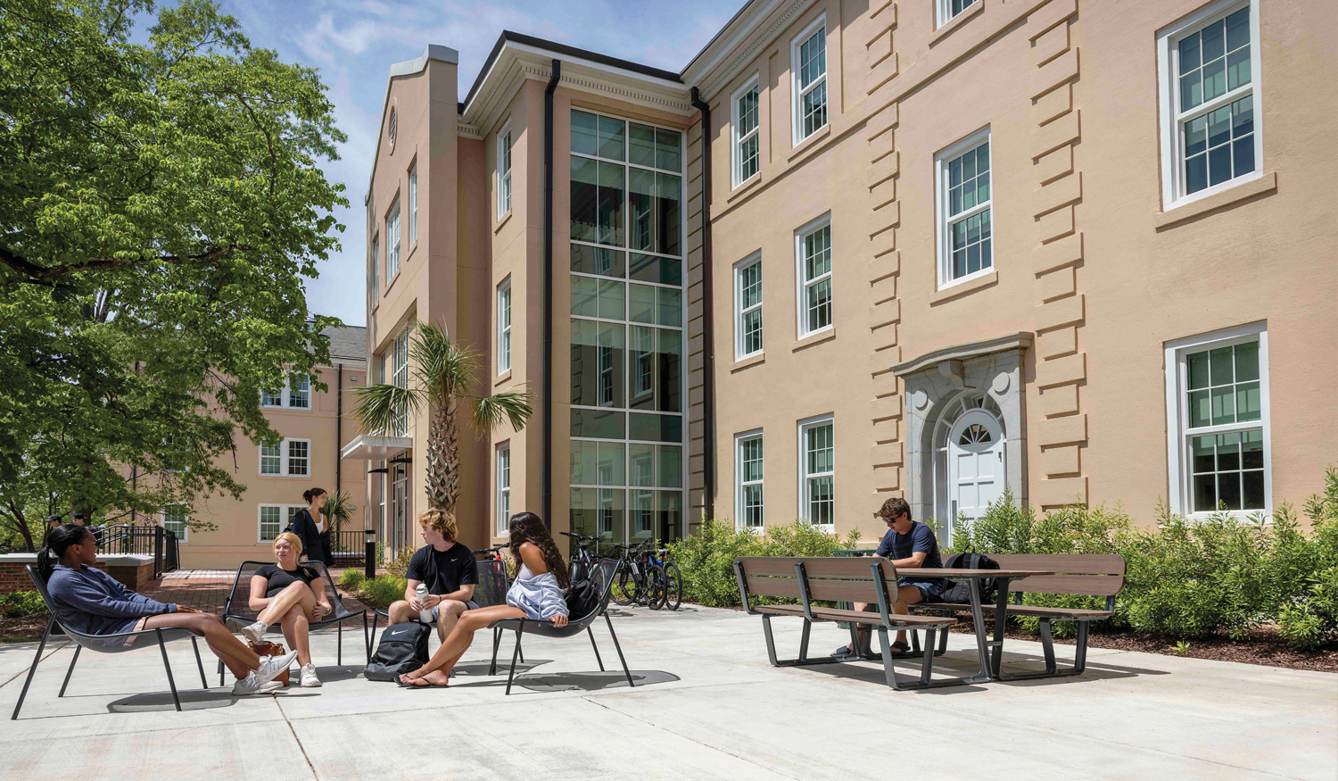 Students relax and socialize in an outdoor courtyard beside a multi-story residence hall. Several students sit in lounge chairs talking, while another studies at a picnic table. Trees, landscaping, bicycles, and large windows create a welcoming campus living environment on a sunny day.