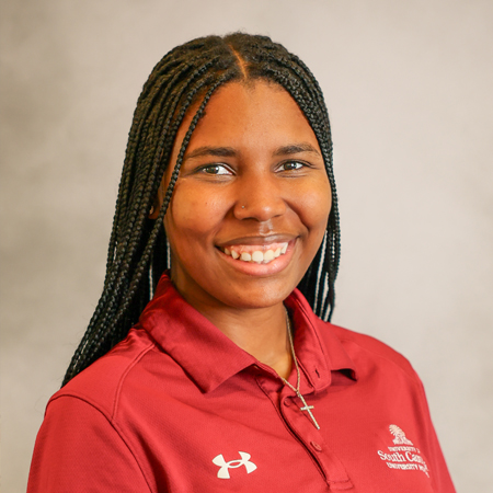 Professional headshot of a smiling person with long black box braids wearing a garnet collared polo shirt with university and Under Armour logos. The person is facing the camera and is wearing a small nose stud and a delicate cross necklace. The photo is set against a soft gray studio background with warm, even lighting, creating a friendly, approachable, and professional appearance.