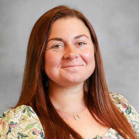 Professional headshot of a smiling person with long auburn hair wearing a light-colored floral blouse with ruffled sleeves and a delicate horseshoe-shaped pendant necklace. The person is facing the camera against a soft gray studio background with even lighting, creating a friendly, approachable, and professional appearance.