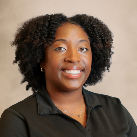 Professional headshot of a smiling person with natural curly black hair wearing a black collared polo shirt, small hoop earrings, and a delicate silver necklace. The person is facing the camera against a soft gray studio background with warm, even lighting, creating a friendly, approachable, and professional appearance.