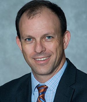 Peter Duffy, smiling, wearing a dark gray suit jacket over a light blue dress shirt with a striped orange and blue tie.