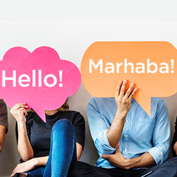 Two people sitting next to each other. One holds up a paper speech bubble that reads "Hello!" while the other has one that says "Marhaba!"