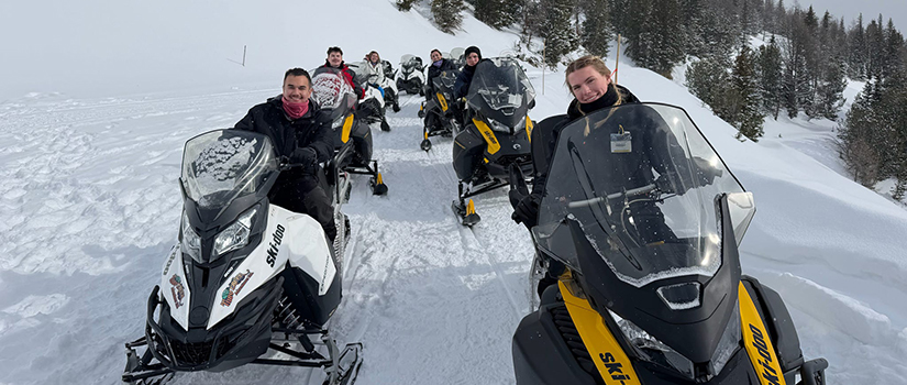 A group of six people ride snowmobiles along a groomed trail through a snowy mountain landscape. Two riders in the foreground smile toward the camera while evergreen trees and snow-covered slopes stretch into the background.