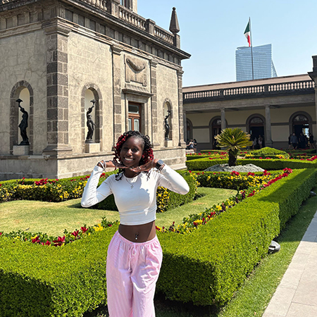 A student poses in a historic courtyard in Mexico City, standing beside manicured hedges and colorful flower beds. The stone architecture of the museum surrounds the garden, with a Mexican flag visible in the background.