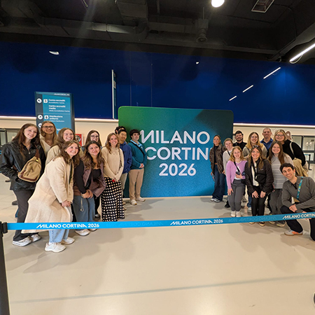 A group of students poses for a photo at a Milano Cortina 2026 venue, gathered around a large sign displaying the Olympic and Paralympic Winter Games branding during an international study experience.
