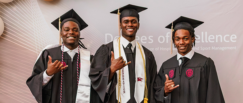 Three University of South Carolina graduates from the College of Hospitality, Retail and Sport Management pose in caps and gowns at the Degrees of Excellence ceremony, smiling and making the “Gamecock” hand sign in front of a branded event backdrop.