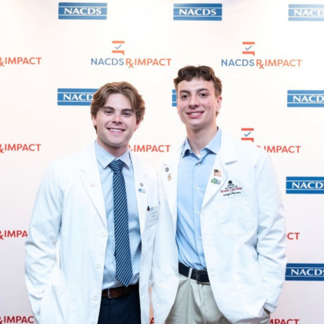 Two male students in white coats in front of a step-and-repeat photo backdrop