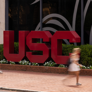 bold garnet USC letters outside of a gray building with students walking by